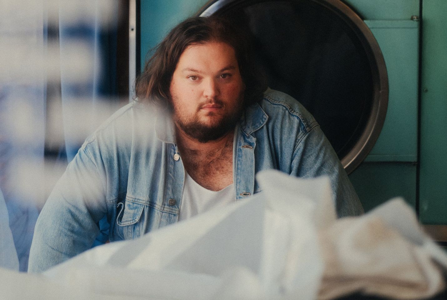 A man with dark hair and a beard wearing a white tshirt and denim shirt